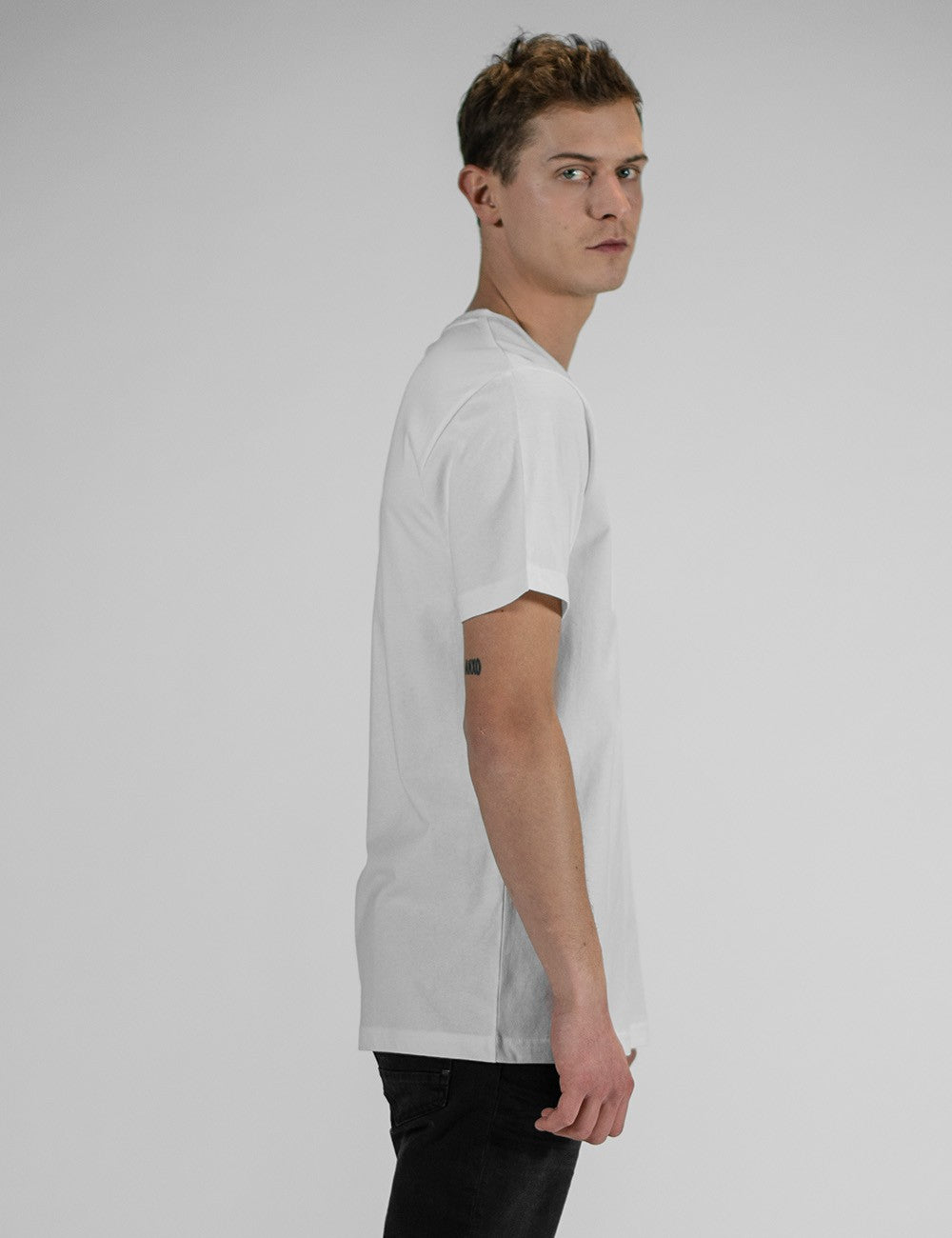 A man in side profile, wearing a plain white t-shirt and black pants, looking back over his shoulder against a grey background.