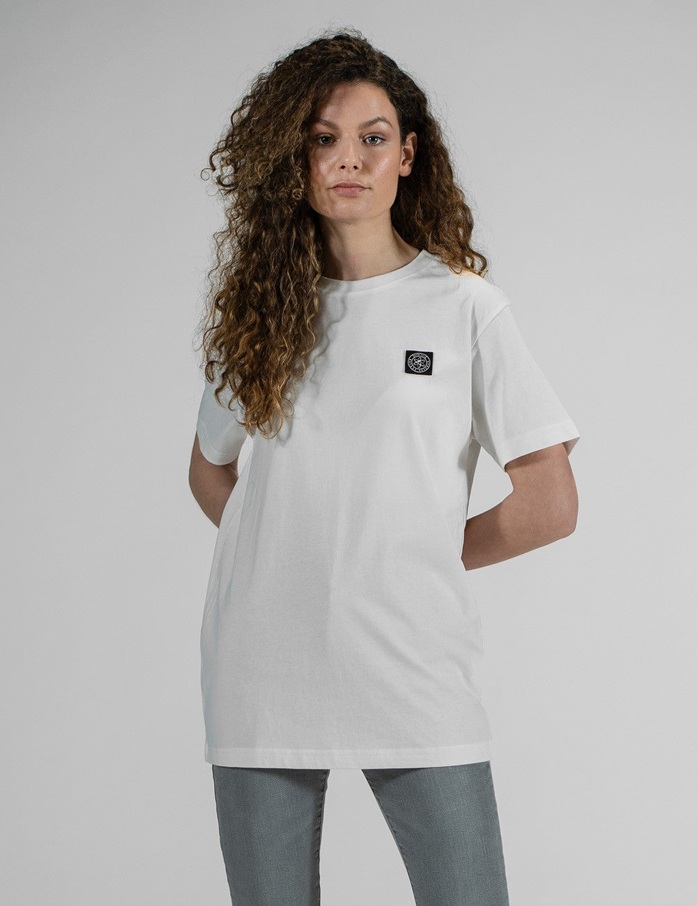 Front pose of a young woman with long, curly brown hair wearing an oversized plain white t-shirt and grey pants against a light grey background.