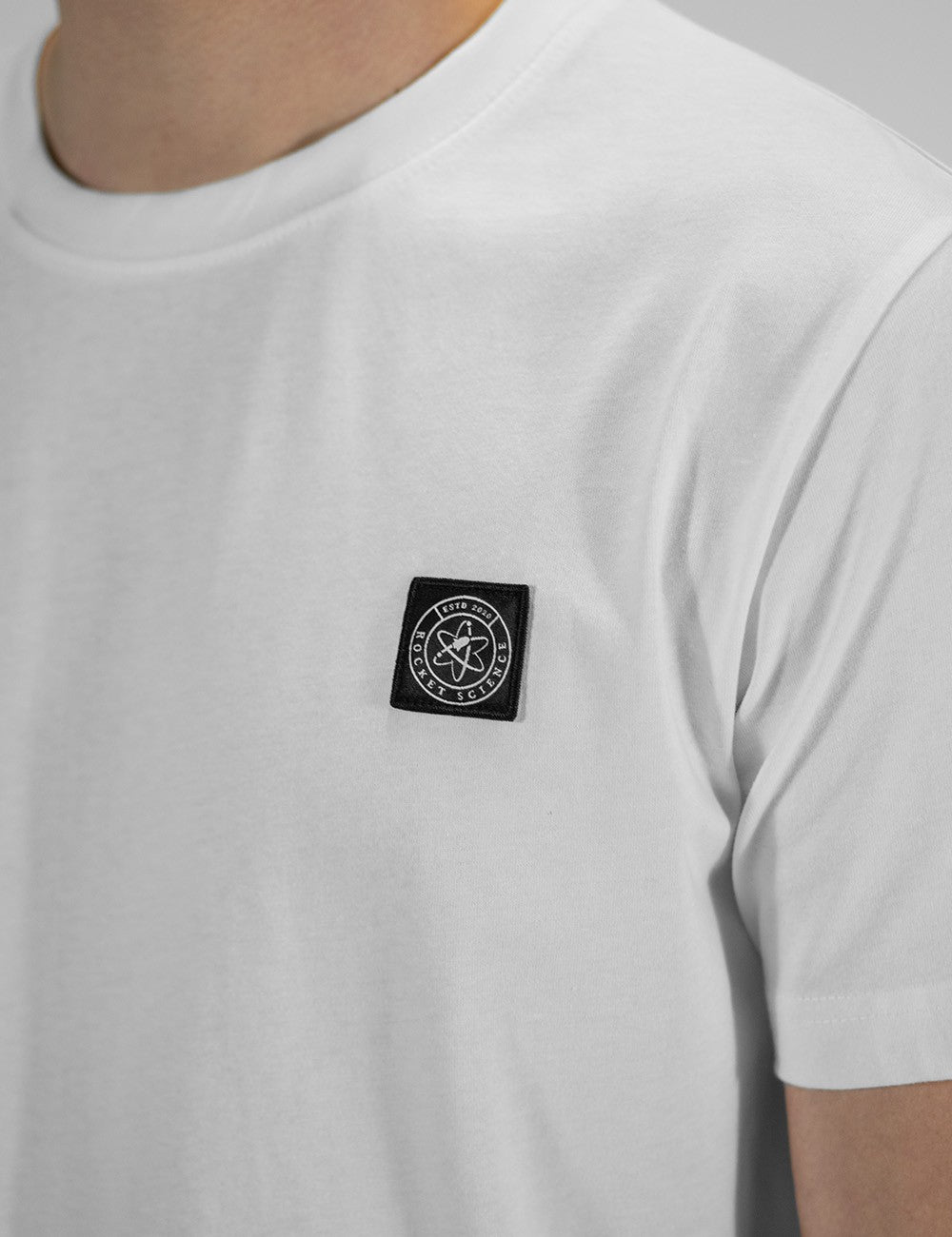 A close-up shot of a person wearing a white, crewneck t-shirt. On the left chest area, there is a small, square black patch with a white circular logo featuring an atom symbol inside it.