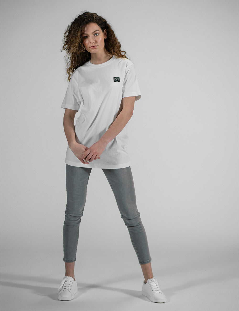 This image shows a young womanwearing a white crew-neck t-shirt with a small chest patch logo and grey skinny jeans.