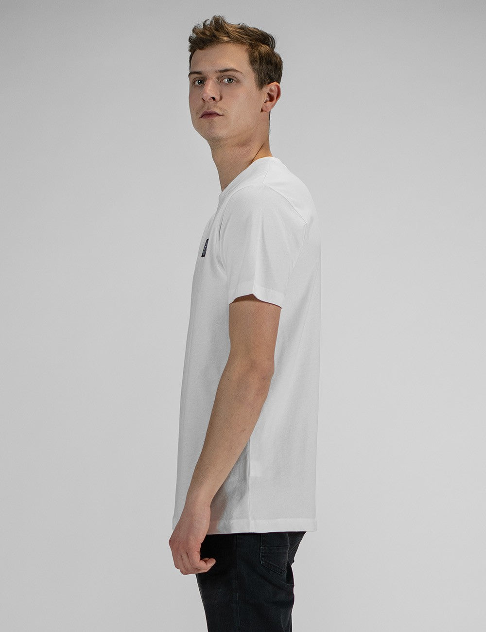 A man in profile, wearing a plain white t-shirt and black pants, looking back over his shoulder.
