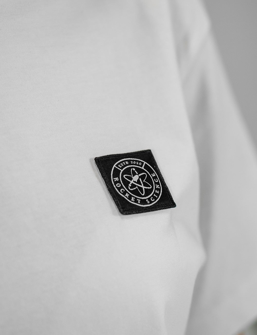 A close-up shot of a small, black, diamond-shaped patch sewn onto a plain white fabric background. The patch features a white circular logo with an atom symbol in the center, surrounded by the words "ROCKET SCIENCE" and "ESTD 2020" in uppercase letters.

