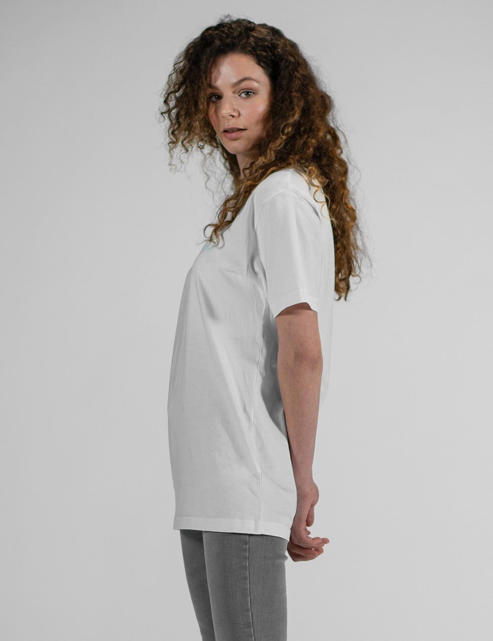 Side profile of a person with long, curly brown hair wearing an oversized plain white t-shirt and grey pants against a light grey background.