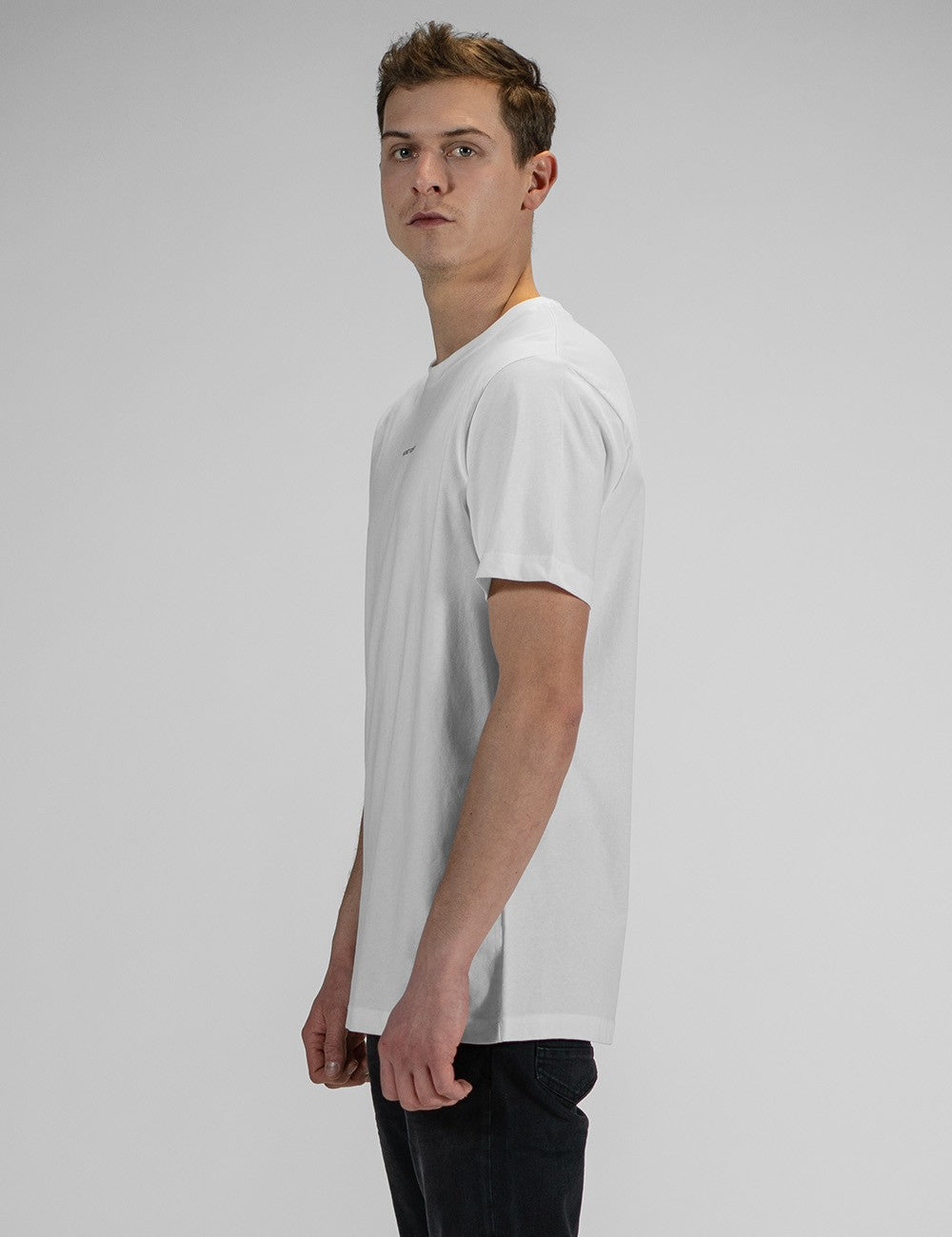 A side profile shot of a young man with short brown hair, looking slightly over his shoulder toward the camera. He is wearing a plain white, short-sleeved crew-neck t-shirt.