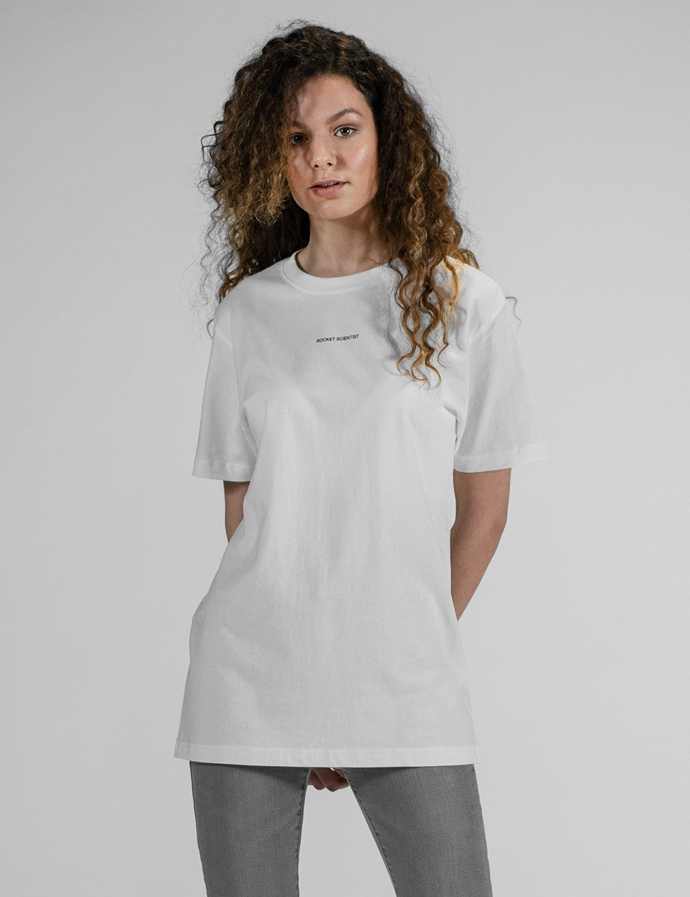 A young woman with long, voluminous, curly brown hair wearing an oversized plain white t-shirt.