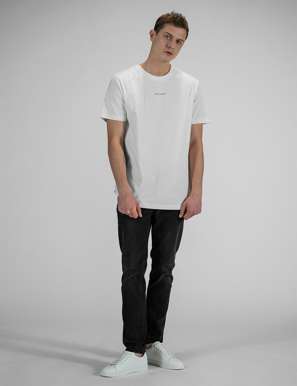 A young man with short brown hair looks directly forward while wearing a plain white, crew-neck, short-sleeve t-shirt that features a tiny line of black text "Rocket Scientist" printed horizontally across the upper chest.