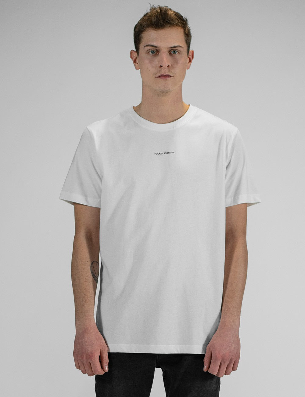 A man with short brown hair stands against a plain grey background, wearing a simple white short-sleeve crewneck t-shirt with a small, minimalist text print "Rocket Scientist" centered on the chest, paired with black pants.
