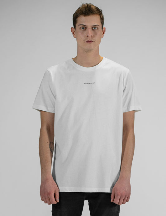 A man with short brown hair stands against a plain grey background, wearing a simple white short-sleeve crewneck t-shirt with a small, minimalist text print "Rocket Scientist" centered on the chest, paired with black pants.