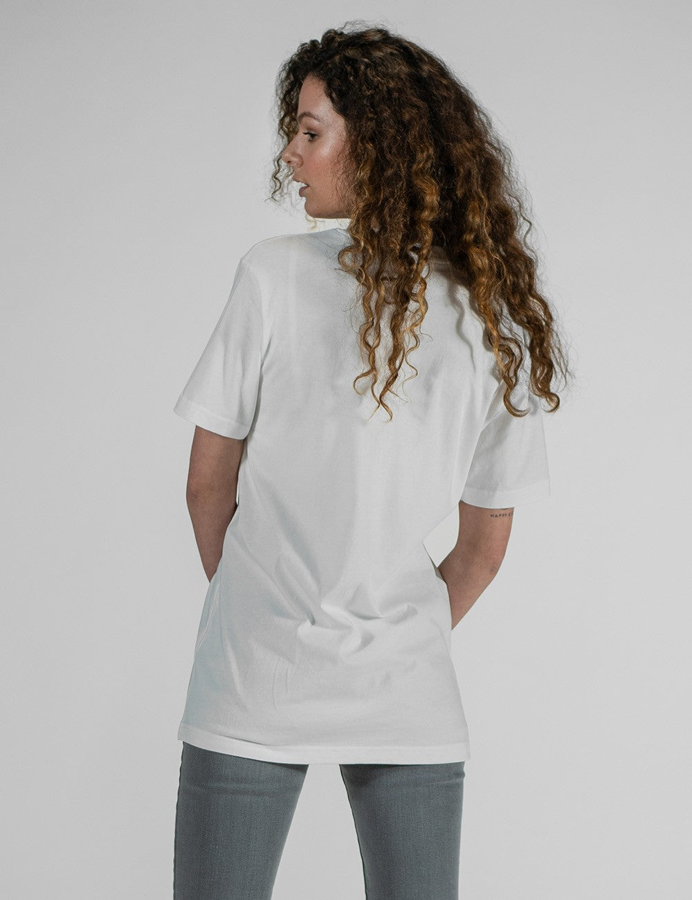 A young woman with long, voluminous, curly brown hair is viewed from the back, looking over her left shoulder to show her profile. She is wearing a relaxed-fit, plain white cotton t-shirt.