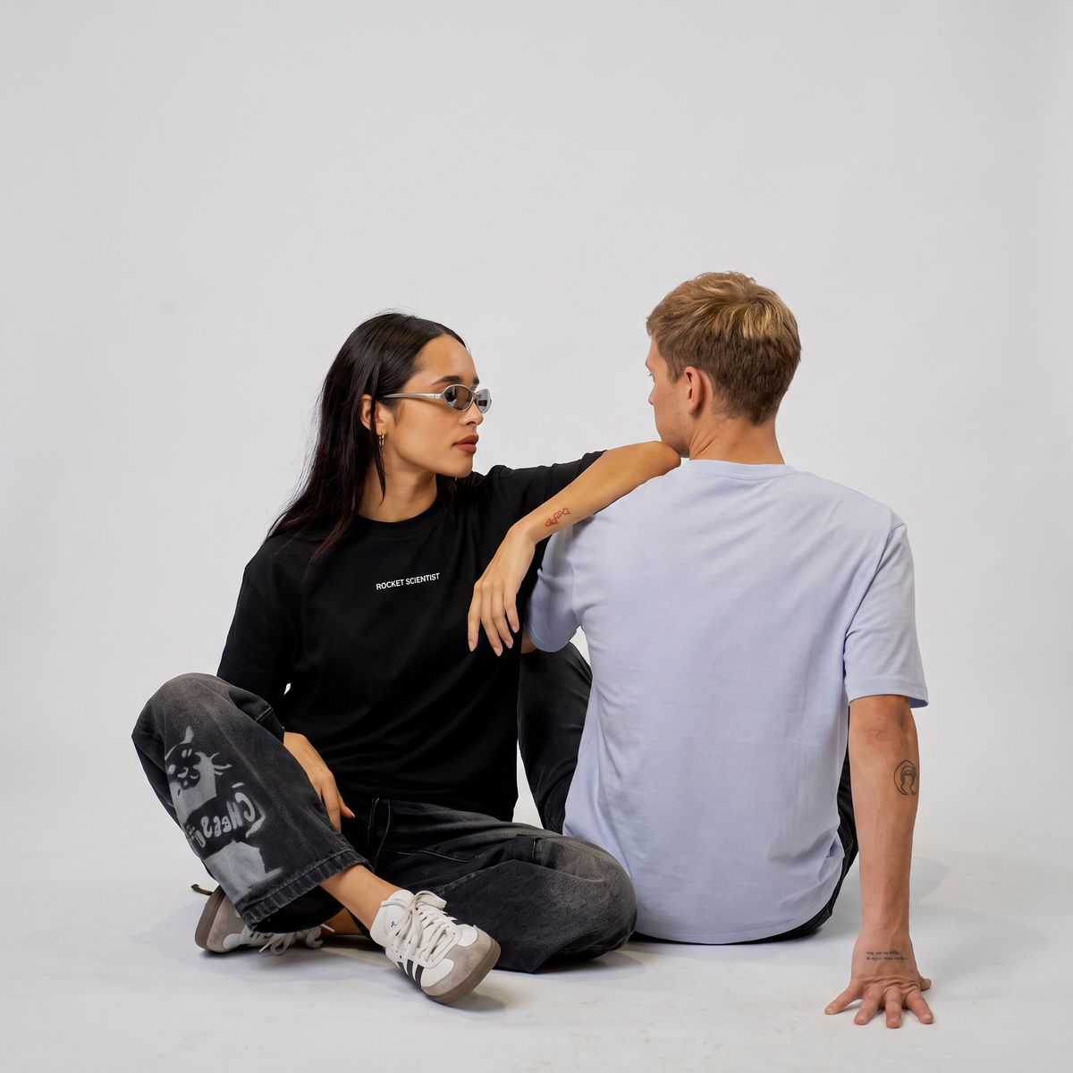A man and a woman sit on a white studio floor facing each other. The woman wears a black t-shirt with small white text on the chest, dark distressed jeans with a graphic print on the left leg, and white sneakers.